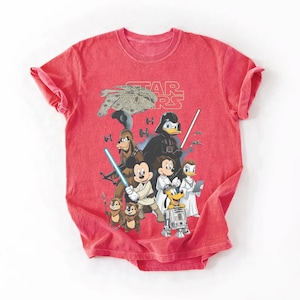 May include: Red t-shirt featuring a graphic of Star Wars characters and Disney characters, including Darth Vader, Mickey Mouse, and Donald Duck. The Star Wars logo is at the top. The shirt has short sleeves and a crew neck.