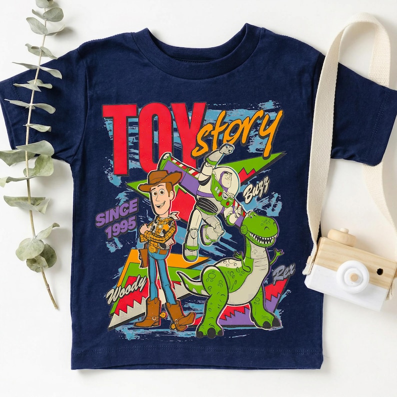 May include: Navy blue t-shirt featuring the Toy Story logo and characters Woody, Buzz Lightyear, and Rex. The shirt includes the text "Since 1995". A fun, colorful graphic tee for fans of the animated movie.