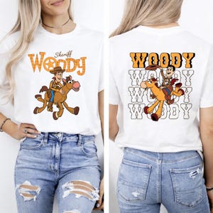 May include: White t-shirt featuring a cartoon graphic of Woody riding a horse. The front of the shirt has the word "Woody" in orange and a sheriff's star. The back of the shirt repeats the Woody graphic and the word "Woody" in orange.