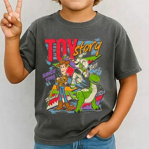 May include: Dark gray t-shirt featuring the words "Toy Story" in red and yellow, with Woody, Buzz Lightyear, and Rex the dinosaur. The shirt has the text "Since 1995" and a colorful graphic design.