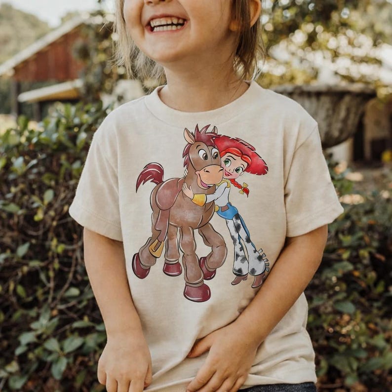 Op de afbeelding: Beige T-shirt met korte mouwen met een cartoonillustratie van een bruin paard dat een cowgirl met rood haar en een rode hoed omhelst. De cowgirl draagt een blauw shirt, een blauwe jeans met witte stippen en een witte riem.