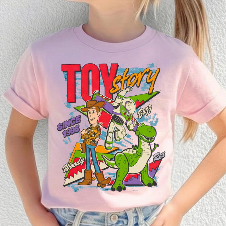 May include: A light pink t-shirt featuring the words "Toy Story" in red and yellow, with the phrase "Since 1995." The shirt displays Woody, Buzz Lightyear, and Rex the dinosaur. The design has a retro, cartoon style.