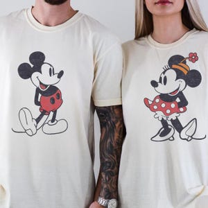 May include: Cream-colored t-shirts featuring Mickey and Minnie Mouse. Mickey is depicted in his classic red shorts, while Minnie wears a red polka-dot skirt and a flower-adorned hat. The cartoon characters are in a vintage style.