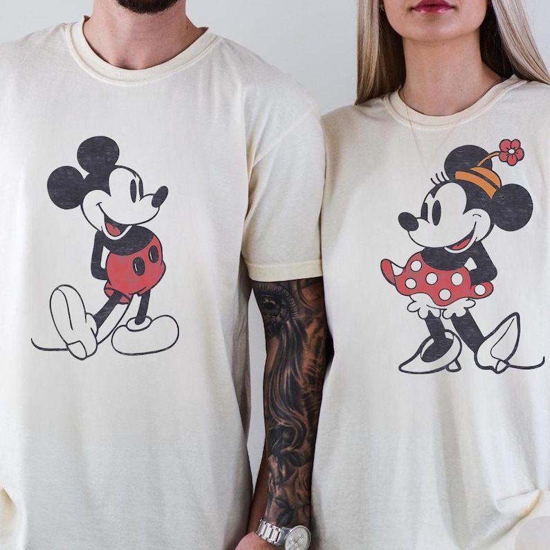 May include: Cream-colored t-shirts featuring Mickey and Minnie Mouse. Mickey is depicted in his classic red shorts, while Minnie wears a red polka-dot skirt and a flower-adorned hat. The cartoon characters are in a vintage style.