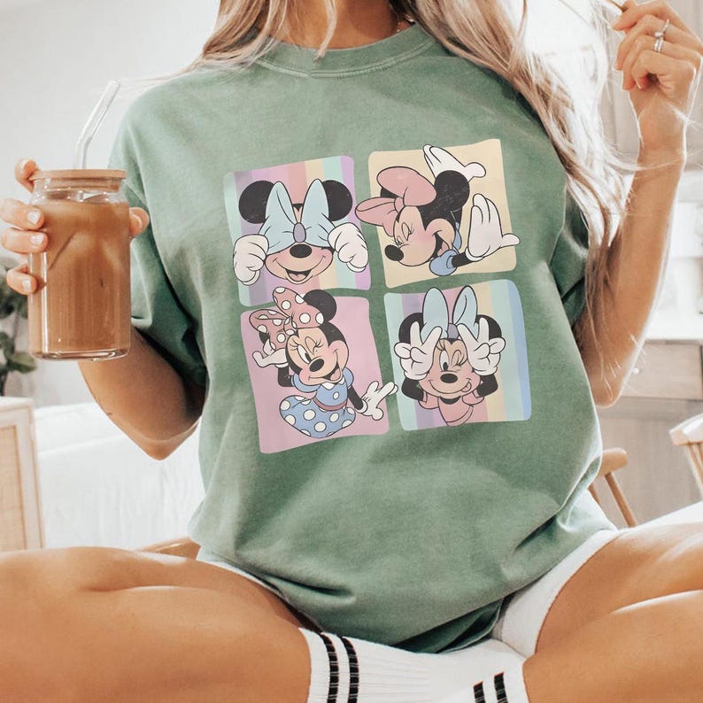 May include: Sage green t-shirt featuring a four-panel graphic of Minnie Mouse in various poses. Each panel has a pastel rainbow background. Minnie is wearing different bows and outfits. The shirt is a relaxed fit.