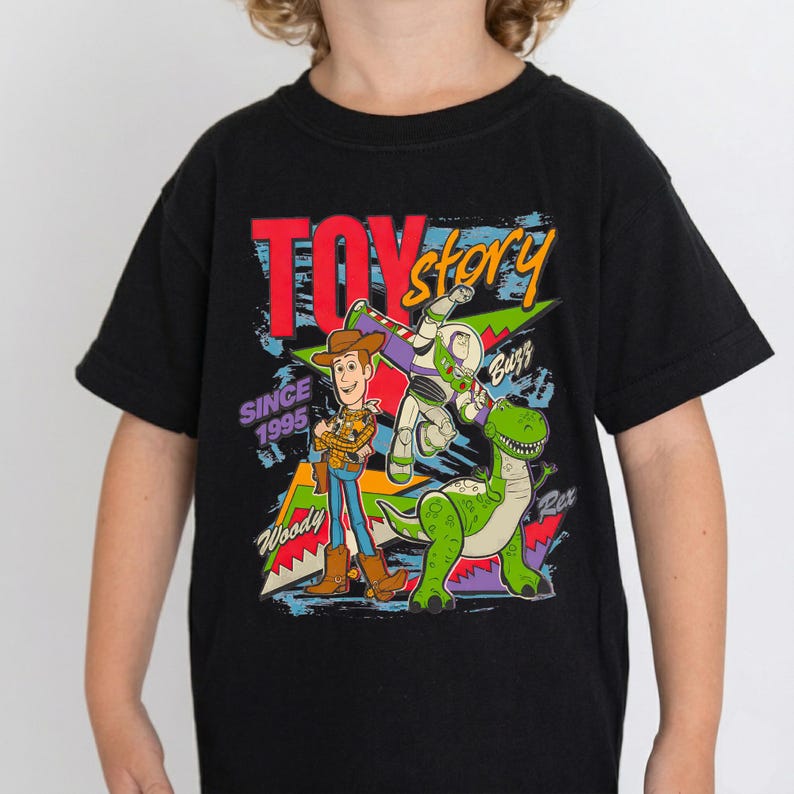 May include: Black t-shirt featuring a colorful graphic of Toy Story characters Woody, Buzz Lightyear, and Rex. The words "Toy Story" and "Since 1995" are also displayed. The shirt is made of a soft material.