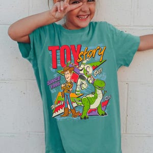 May include: Teal-colored t-shirt featuring the words "Toy Story" in red and yellow, with Woody, Buzz Lightyear, and Rex graphics. The shirt also includes the text "Since 1995".