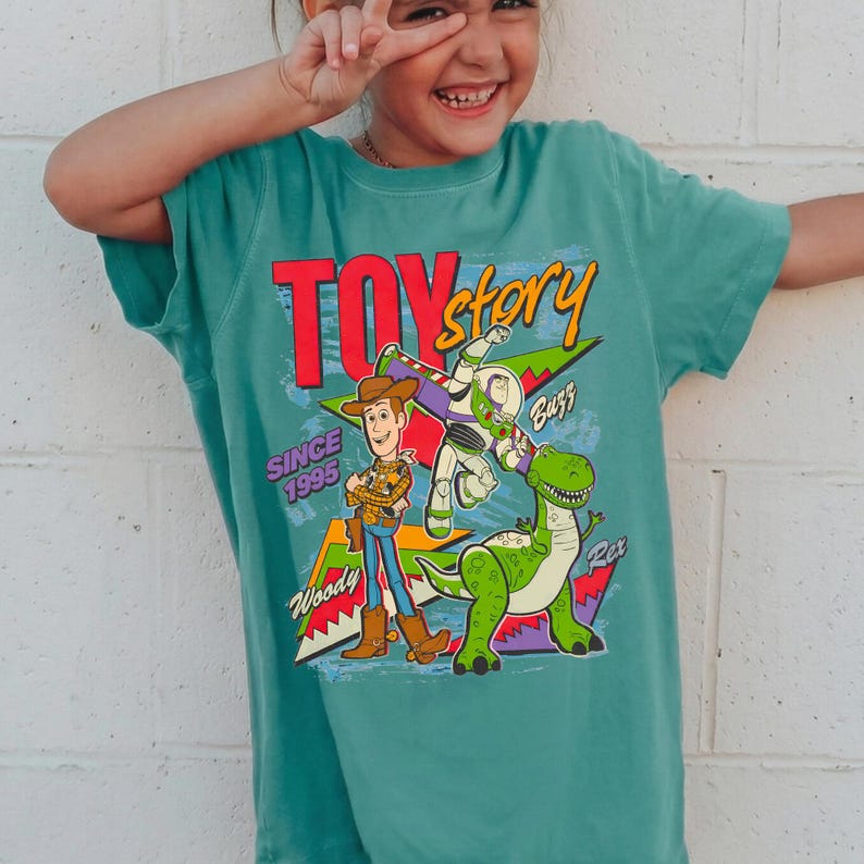 May include: Teal-colored t-shirt featuring the words "Toy Story" in red and yellow, with Woody, Buzz Lightyear, and Rex graphics. The shirt also includes the text "Since 1995".
