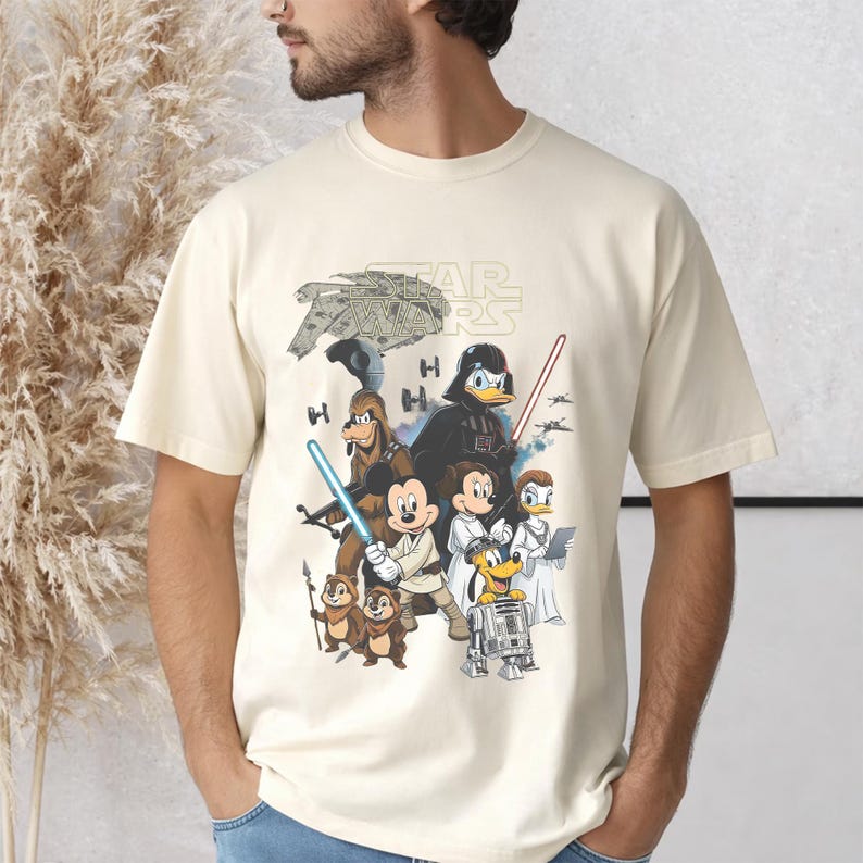 May include: Cream-colored t-shirt featuring a Star Wars graphic with Disney characters. The design includes Darth Vader as Donald Duck, Mickey Mouse as Luke Skywalker, and other characters from both franchises. The Star Wars logo is at the top.