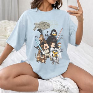 May include: Light blue t-shirt featuring a graphic of Star Wars characters, including Darth Vader, Mickey Mouse, and Donald Duck. The Star Wars logo is at the top. The shirt has short sleeves and a crew neck.