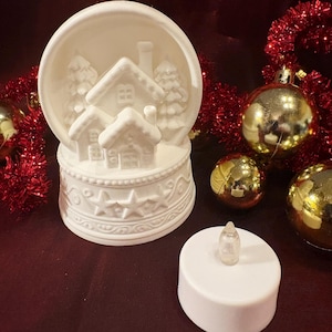 May include: A white ceramic Christmas snow globe featuring a village scene with houses and trees. The base is decorated with stars and swirls. A white LED candle sits in front of the globe, with gold ornaments and red tinsel in the background.