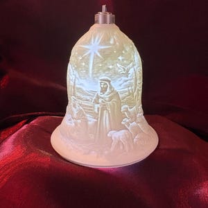 May include: A white, bell-shaped Christmas ornament with a religious scene carved into the surface. The design includes a star, figures, and animals. The ornament has a silver metal top and is lit from within.