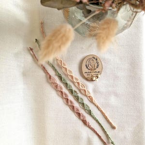 Peut inclure: Quatre bracelets en macramé dans les tons de rose, vert et beige, disposés en diagonale sur une surface blanche. Un petit disque en bois avec un logo et du texte est également présent. Des fleurs séchées et des éléments décoratifs sont en arrière-plan.
