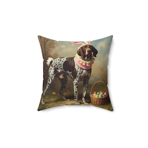 May include: Square throw pillow featuring a brown and white spotted dog wearing bunny ears and a pink and white ruffled collar. A wicker basket filled with colorful Easter eggs sits nearby, with eggs scattered on the grass.