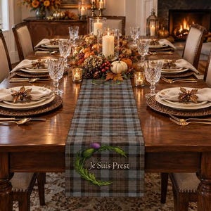 May include: A rectangular table runner featuring a grey and brown plaid pattern. The runner displays a green thistle graphic with the text Je Suis Prest printed in a serif font. The fabric is centered on a wooden dining table with place settings.