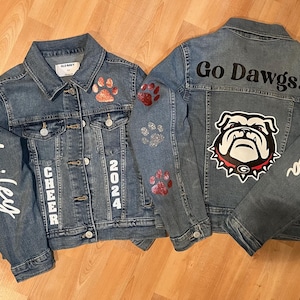 May include: Two denim jackets with custom designs. One jacket features the name "Hailey", "CHEER", and "2024" in white lettering, along with paw print accents. The other jacket has "Go Dawgs!" and a bulldog graphic.