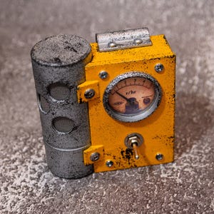 Puede incluir: Un dispositivo rectangular amarillo con acabado desgastado, que presenta un medidor analógico circular etiquetado como r/hr, un interruptor de palanca metálico y un componente lateral gris cilíndrico con aberturas de malla, fijado con tornillos plateados.