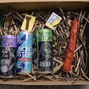 May include: A gift box with three energy drink cans, a sausage, a bolt, and a matchbox. The cans are labeled "NON STOP" with different designs, including a figure wearing a gas mask. The sausage is red.