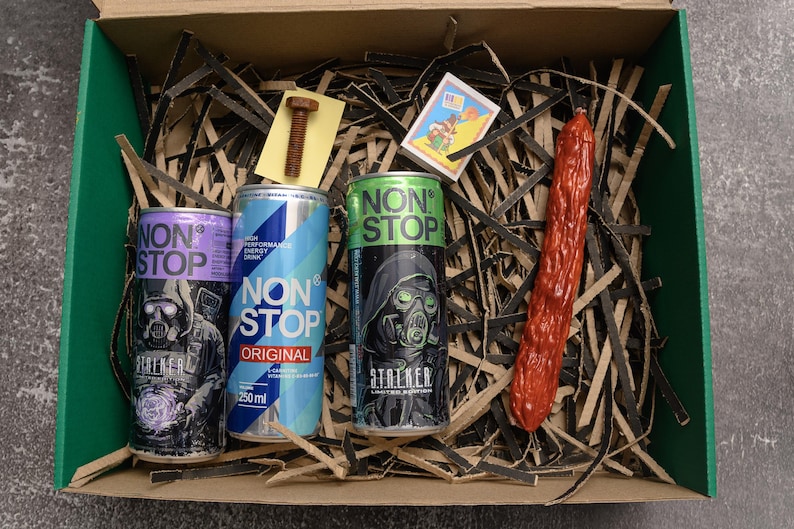 May include: A gift box with three energy drink cans, a sausage, a bolt, and a matchbox. The cans are labeled "NON STOP" with different designs, including a figure wearing a gas mask. The sausage is red.