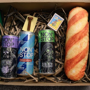 May include: A gift box contains three cans of "NON STOP" energy drink, a bolt, a matchbox, and a long bread-like item with red stripes. The energy drink cans are purple, blue, and green, each with a different design. The box is green.