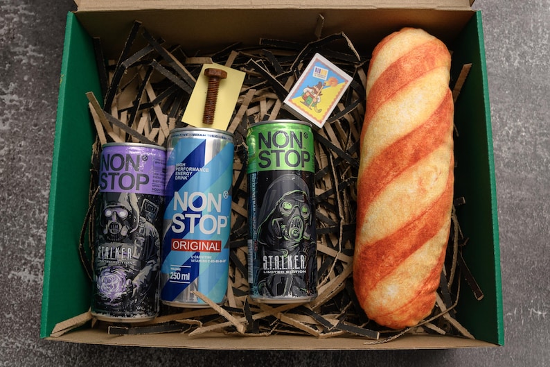 May include: A gift box contains three cans of "NON STOP" energy drink, a bolt, a matchbox, and a long bread-like item with red stripes. The energy drink cans are purple, blue, and green, each with a different design. The box is green.