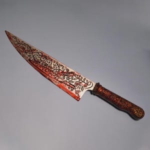 Puede incluir: Un cuchillo grande y ornamentado con una hoja blanca con un diseño floral y detalles en rojo. El mango es de color marrón oscuro con detalles intrincados. El cuchillo es probablemente para fines decorativos.