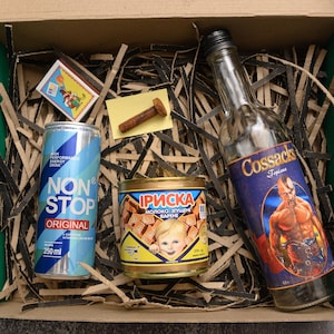 May include: A gift box with a variety of items. Includes a blue and white "NON STOP ORIGINAL" energy drink can, a bottle of "Cossacks" liquor, a can of "Iриска" candy, a box of matches, and a bolt. The items are arranged on shredded paper.