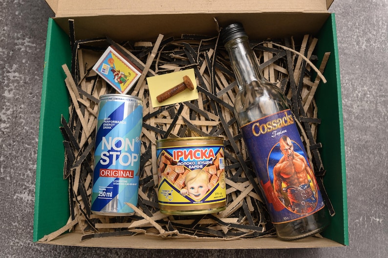 May include: A gift box with a variety of items. Includes a blue and white "NON STOP ORIGINAL" energy drink can, a bottle of "Cossacks" liquor, a can of "Iриска" candy, a box of matches, and a bolt. The items are arranged on shredded paper.