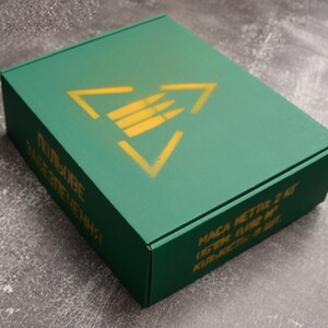 May include: A green rectangular box with a yellow triangle and bullet symbol on the top. The box has text in a foreign language on the side. The box is on a gray surface.