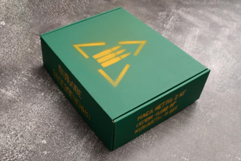 May include: A green rectangular box with a yellow triangle and bullet symbol on the top. The box has text in a foreign language on the side. The box is on a gray surface.