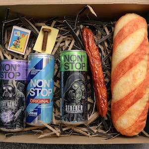 May include: A gift box contains three "NON STOP" energy drink cans, a sausage, a loaf of bread, a small matchbox, and a bolt. The cans are purple, blue, and green, each with a different graphic. The sausage is red, and the bread is striped.