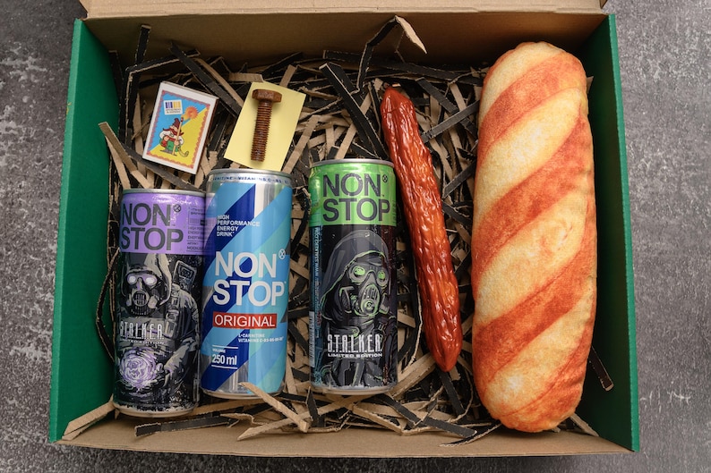 May include: A gift box contains three "NON STOP" energy drink cans, a sausage, a loaf of bread, a small matchbox, and a bolt. The cans are purple, blue, and green, each with a different graphic. The sausage is red, and the bread is striped.