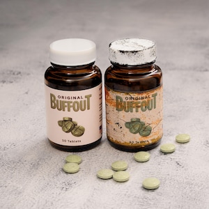 May include: Two amber glass bottles of Buffout tablets with white caps. The left bottle has a clean cream label, while the right bottle has a distressed, weathered label. Both feature the text Original Buffout and 50 Tablets, with loose green tablets scattered around.