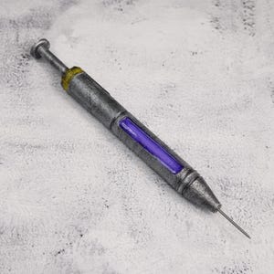 Puede incluir: Una jeringa de utilería cilíndrica con un acabado metálico plateado desgastado, que presenta un anillo dorado cerca del extremo del émbolo y una ventana rectangular central que muestra un inserto de líquido de color púrpura vibrante. Una aguja fina sobresale de la punta.