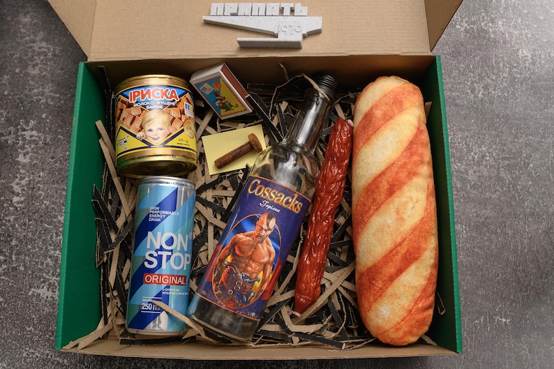 May include: A gift box filled with various food and beverage items. Includes a can of candy, a can of energy drink, a bottle of Cossacks vodka, a sausage, a loaf of bread, and a box of matches. The box is lined with shredded paper.