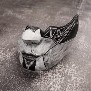 Puede incluir: Una máscara abstracta en blanco y negro hecha a mano. La máscara presenta diseños geométricos y una superficie texturizada. El diseño incluye triángulos y líneas, con una forma de boca estilizada. La máscara es probablemente de cerámica o un material similar.