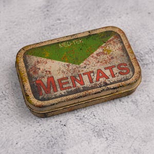 May include: Rectangular metal tin with a distressed, vintage appearance. The top features a green and white design with the word "MENTATS" in red lettering. The text "MED-TEK" is above the design. The tin has a rusty, aged look.