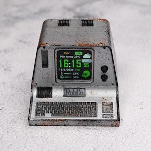 May include: A weathered, metallic gray desktop computer terminal with rust-like accents. The screen displays digital text including 'Kyiv', '16:15', '18/6/2026', and 'RobCo' branding. It features a built-in keyboard, two control knobs, and vents.