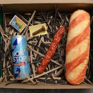May include: A box with a green interior contains a can of "NON STOP ORIGINAL" energy drink, a matchbox, a bolt, a red sausage, and a bread loaf with orange stripes. The items rest on shredded paper.