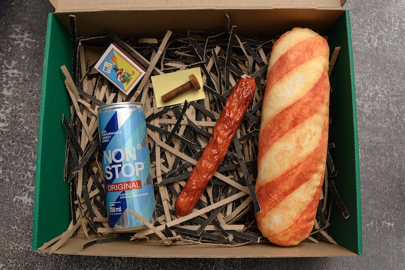 May include: A box with a green interior contains a can of "NON STOP ORIGINAL" energy drink, a matchbox, a bolt, a red sausage, and a bread loaf with orange stripes. The items rest on shredded paper.
