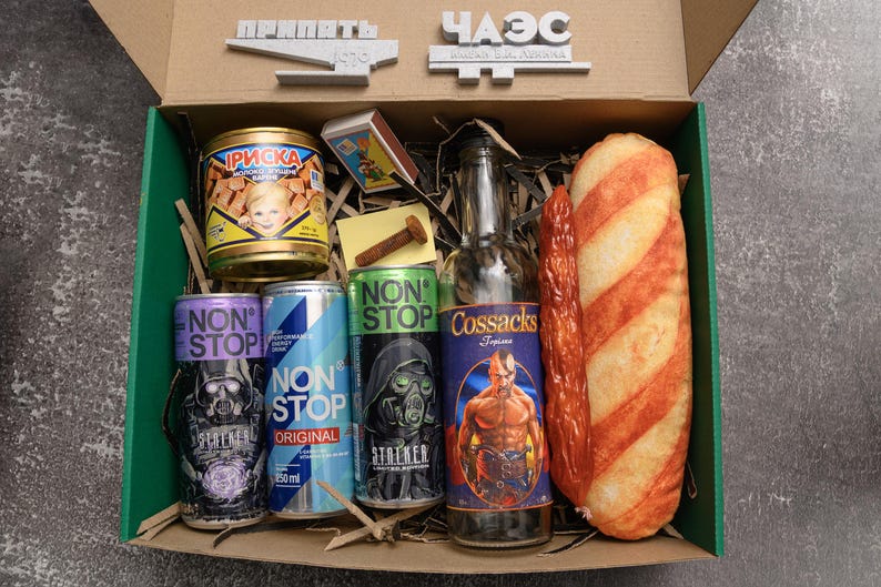 May include: A gift box filled with various items, including canned goods, energy drinks, a bottle, and a loaf of bread. The box contains cans of "NON STOP" and "Cossacks" brand items. The box also has the text "PRIPIAT" and "CHAE" on the top.