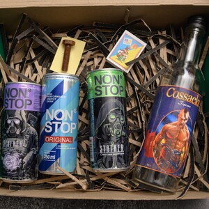 May include: A gift box containing energy drinks and a bottle of liquor. The energy drinks are in purple, blue, and green cans, with the text "NON STOP" and "STREKER". The liquor bottle has a label with a muscular man.