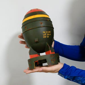 Könnte beinhalten: Ein Bombenmodell in Olivgrün mit roter Nasenkegelspitze und einem gelben horizontalen Streifen. Das Gehäuse zeigt sichtbare Schrauben und den Aufdruck MK-211 NUC LOT 23U-A UNIT 07. Es verfügt über eine kreisförmige Basis mit vier Stabilisierungsflossen.