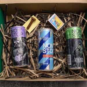 May include: A cardboard box with green edges contains three energy drink cans. The cans are labeled "NON STOP" and feature graphics of a figure in a gas mask. The box also holds shredded paper, a bolt, and a matchbox.