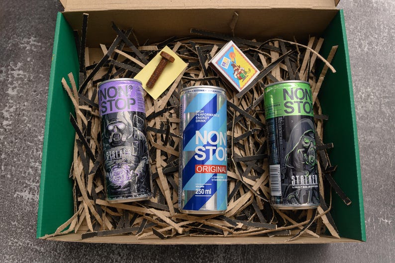 May include: A cardboard box with green edges contains three energy drink cans. The cans are labeled "NON STOP" and feature graphics of a figure in a gas mask. The box also holds shredded paper, a bolt, and a matchbox.