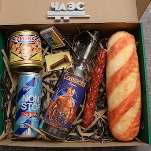 May include: A gift box filled with various food and beverage items. Includes a can of food with a child's image, a can of blue energy drink, a bottle of Cossacks vodka, a sausage, a loaf of bread, and a box of matches.