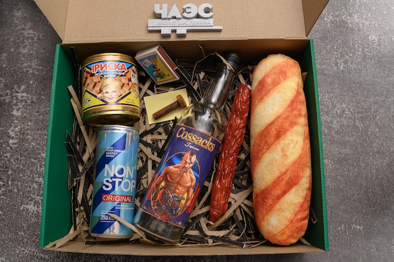 May include: A gift box filled with various food and beverage items. Includes a can of food with a child's image, a can of blue energy drink, a bottle of Cossacks vodka, a sausage, a loaf of bread, and a box of matches.