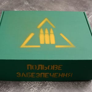 May include: A green cardboard box with a yellow graphic of three bullets inside a triangle. The box has text in a foreign language on the front. The box is rectangular and has a matte finish.