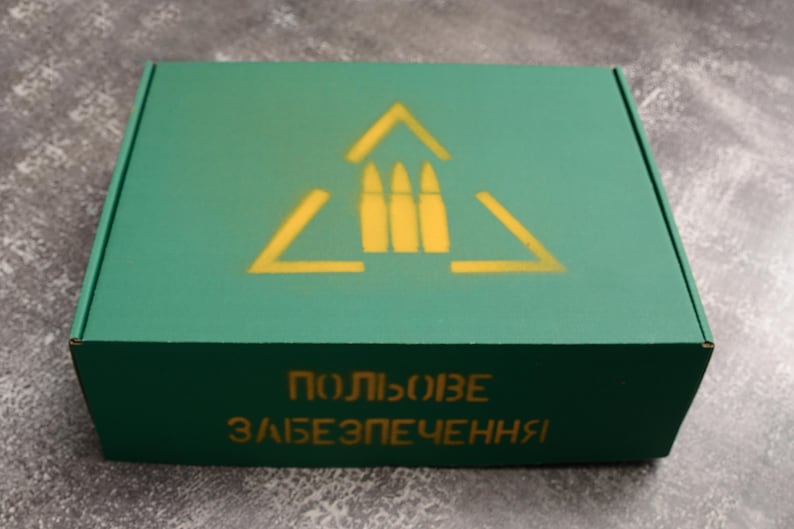 May include: A green cardboard box with a yellow graphic of three bullets inside a triangle. The box has text in a foreign language on the front. The box is rectangular and has a matte finish.