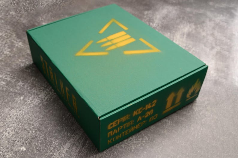 May include: A green rectangular box with a yellow triangle and arrow design on the lid. The box has text in yellow, including the word "STALKER" on the side. The box also has other text and symbols.